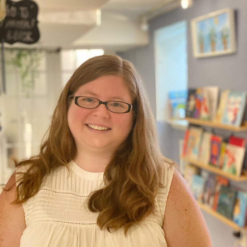 Heather Jenkins standing in Wise Wonder with a wall of books in the background. She is wearing glasses and smiling in a white tank top shirt.