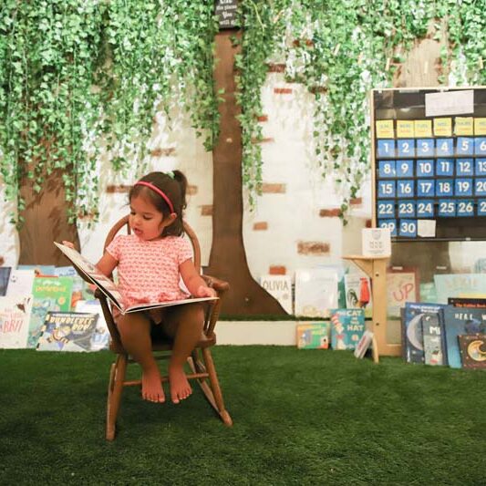 Young girl reading a book in a classroom with astroturf and trees crafted on the walls