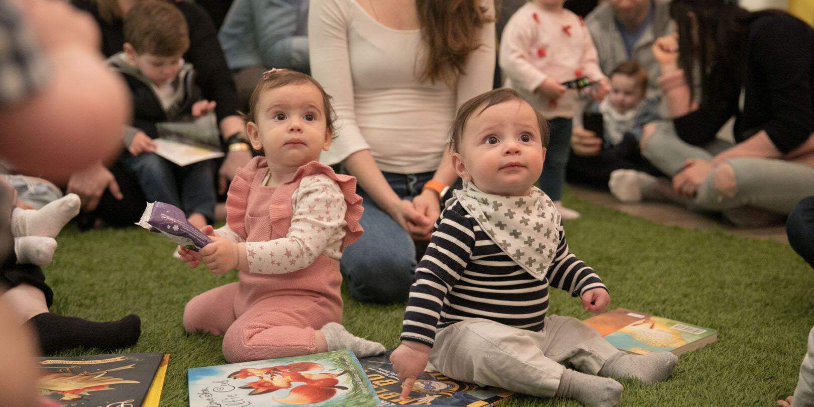 Two toddlers sitting on the floor with books, looking up at a teacher reading to them. Their mothers are sitting behind them.